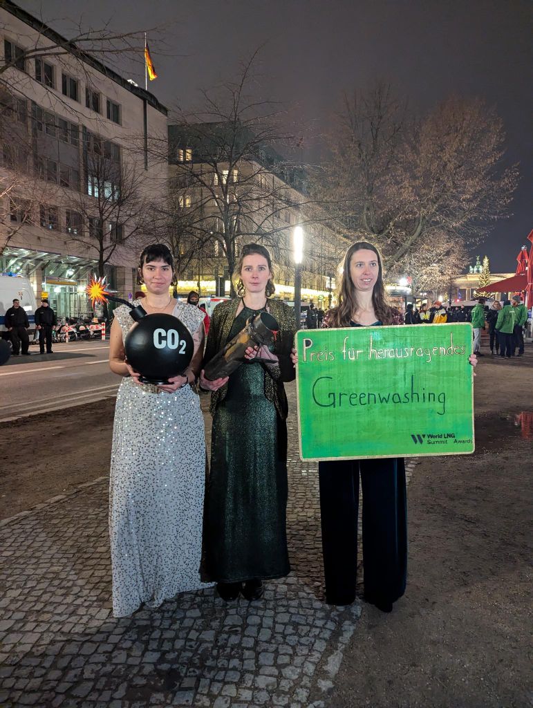 Drei Personen in Abendkleidern mit einer CO2-Bombe, einer blutigen Pipeline und einem grünen Schild "Preis für herausragendes Greenwashing"